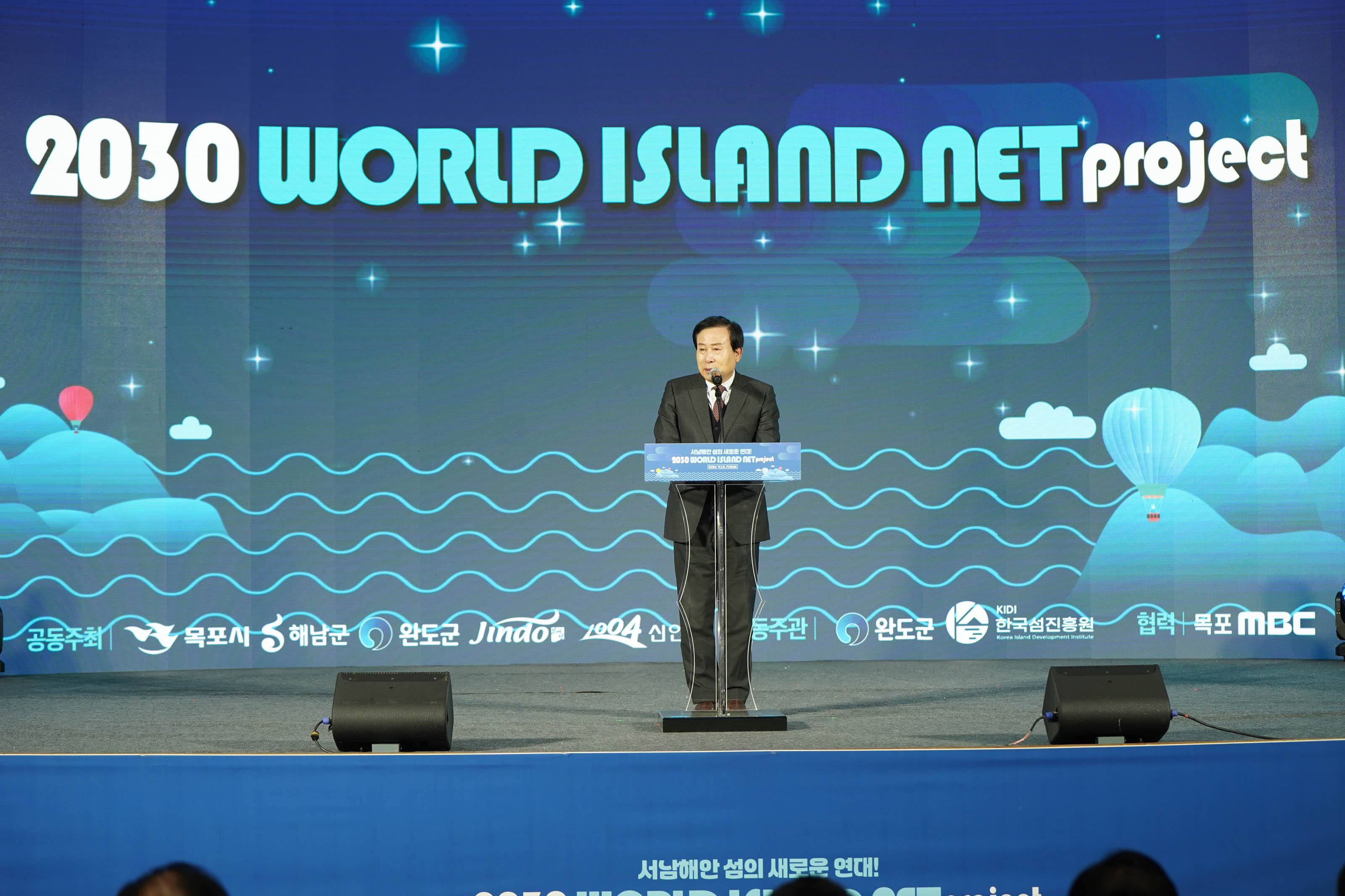 ‘2030 WORLD ISLAND NET project’라는 문구가 적힌 파란색 배경 무대 앞 단상에서 박홍률 목포시장이 마이크를 들고 연설을 하고 있는 모습. 배경에는 별, 구름, 열기구, 파도 무늬가 애니메이션처럼 디자인되어 있다.