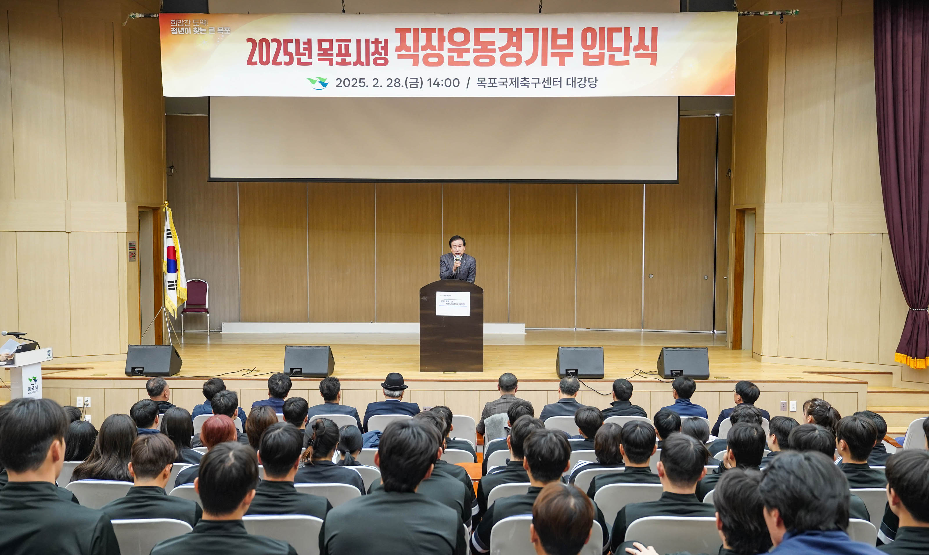 무대 위에 ‘2025년 목포시청 직장운동경기부 입단식’, ‘2025. 2. 28.(금) 14:00 / 목포국제축구센터 대강당’이라고 적힌 큰 현수막이 걸려 있음. 연단에 서서 마이크를 들고 발언 중인 박홍률 목포시장이 보이고, 관객석에는 정장을 입은 사람들과 유니폼을 입은 체육인들이 앉아 있음. 좌측에는 태극기와 목포시 로고가 있는 연단이 있음.
