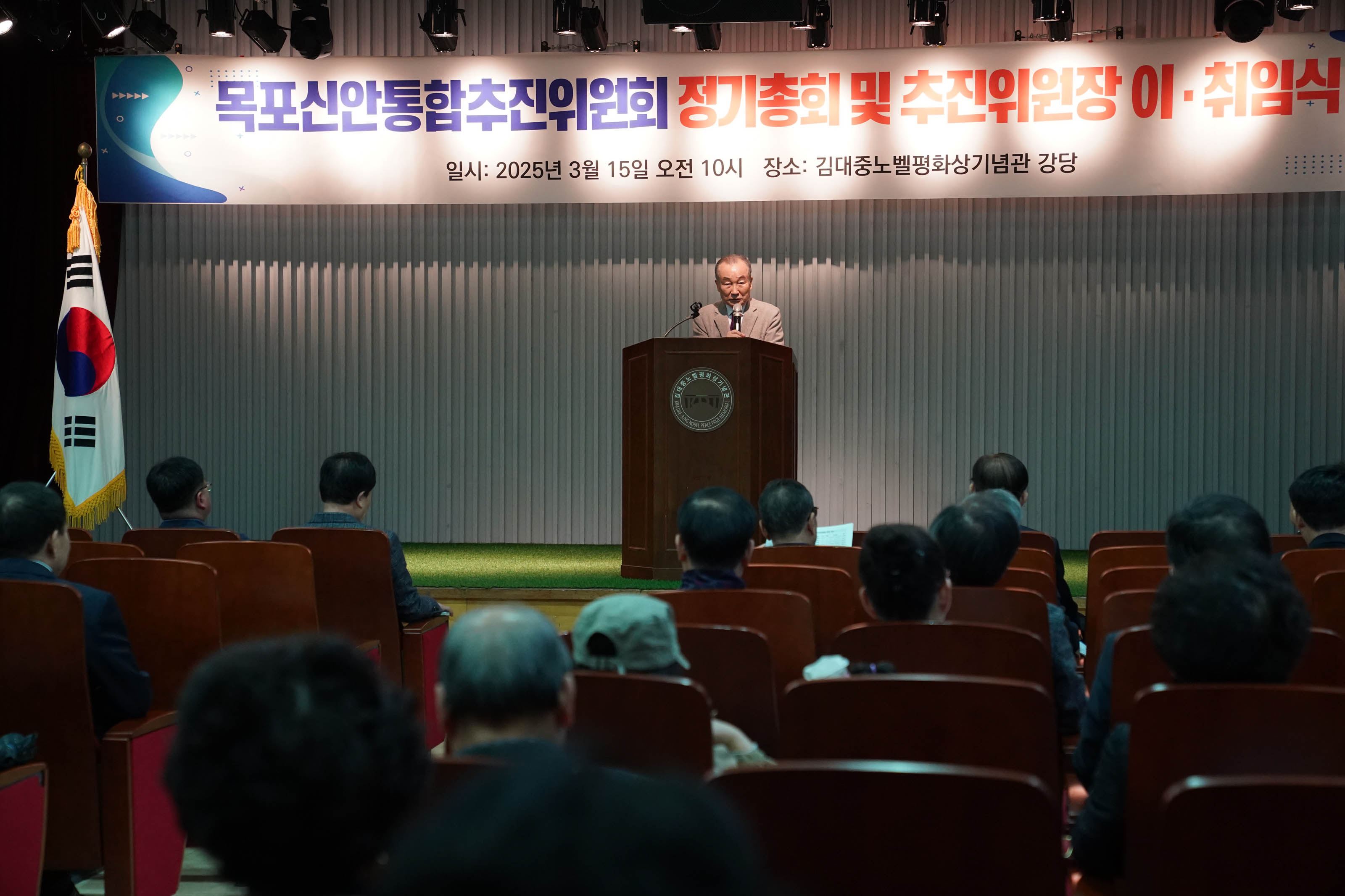 목포신안통합추진위원회 정기총회 및 추진위원장 이·취임식에서 남성이 연단에서 발언하고 있다. 배경 배너에는 '목포신안통합추진위원회 정기총회 및 추진위원장 이·취임식'이라는 문구가 적혀 있고, 그 아래에는 '일시: 2025년 3월 15일 오전 10시'와 '장소: 김대중노벨평화상기념관 강당'이 적혀 있다. 청중들이 자리에 앉아 있으며 발언을 듣고 있다.