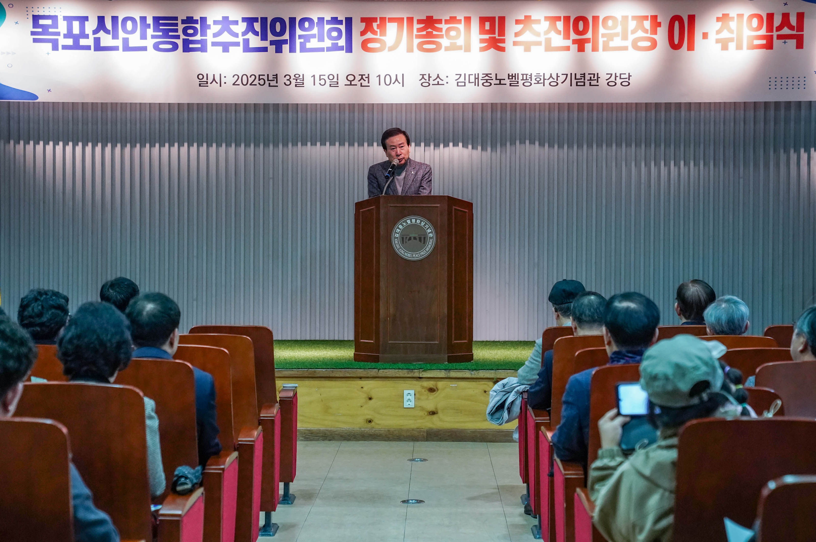 목포신안통합추진위원회 정기총회 및 추진위원장 이·취임식에서 박홍률 목포시장이 연단에서 발언하고 있다. 청중들은 자리에 앉아 이 발언을 듣고 있으며, 배경에는 '목포신안통합추진위원회 정기총회 및 추진위원장 이·취임식'이라는 문구와 '일시: 2025년 3월 15일 오전 10시', '장소: 김대중노벨평화상기념관 강당'이 적혀 있다.