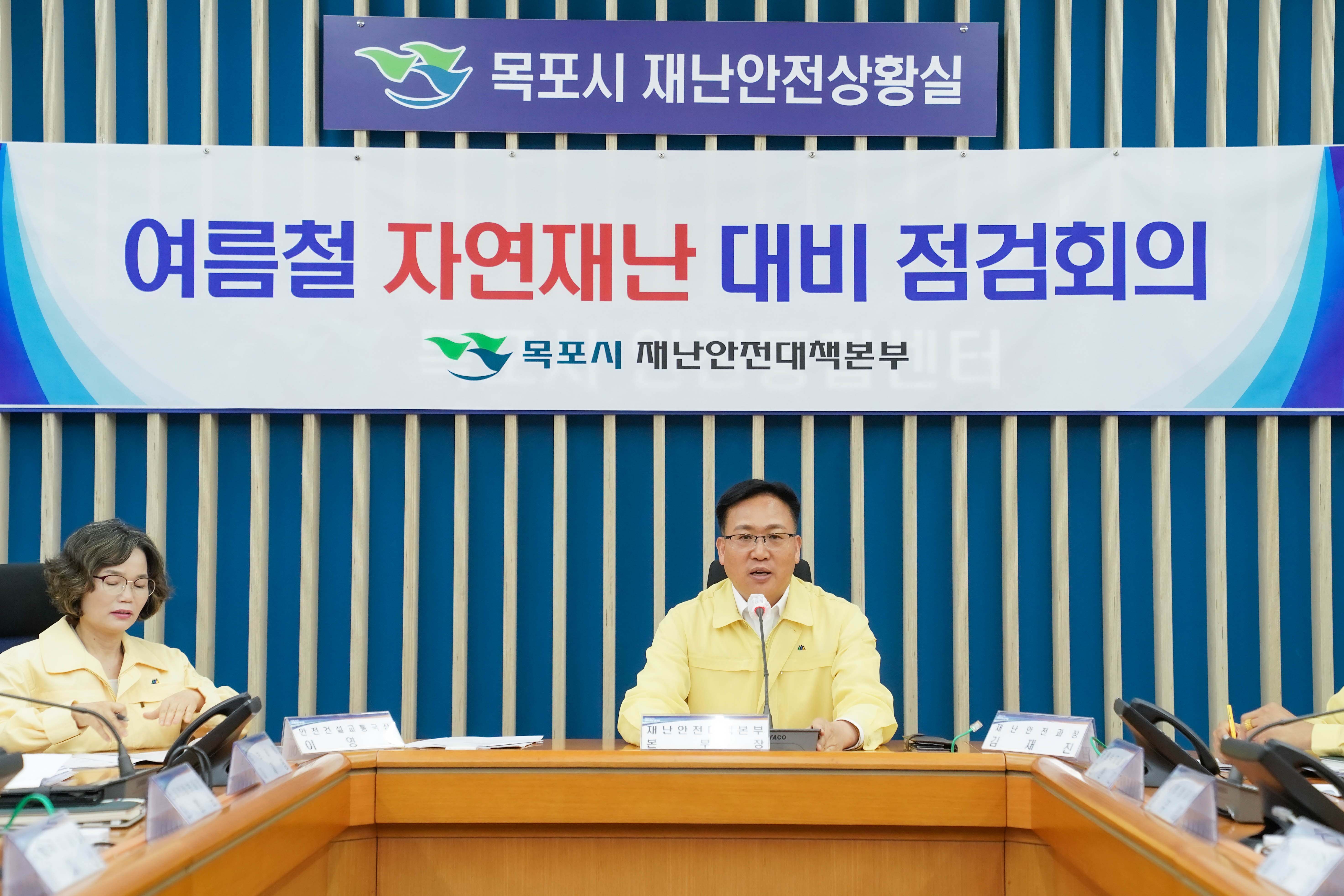 목포시 재난안전상황실에서 열린 '여름철 자연재난 대비 점검회의' 장면. 배경 현수막에는 '목포시 재난안전대책본부' 문구와 로고가 있으며, 노란색 민방위복을 입은 관계자가 발언하고 있습니다.