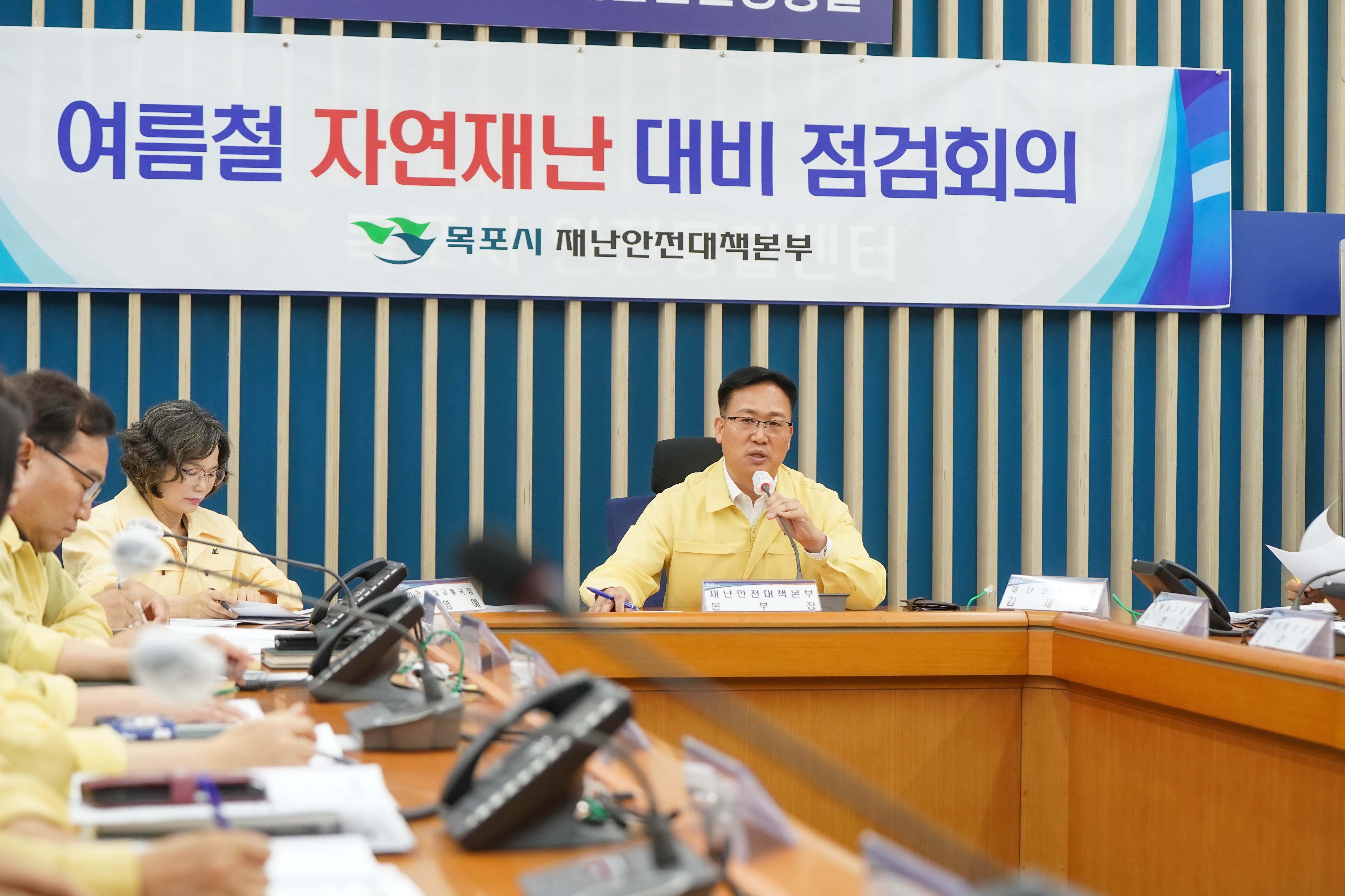 '여름철 자연재난 대비 점검회의' 현수막을 배경으로 노란색 민방위복을 입은 관계자가 마이크를 잡고 발언하는 근접 사진. 테이블 위에는 명패와 회의 자료가 놓여 있습니다.