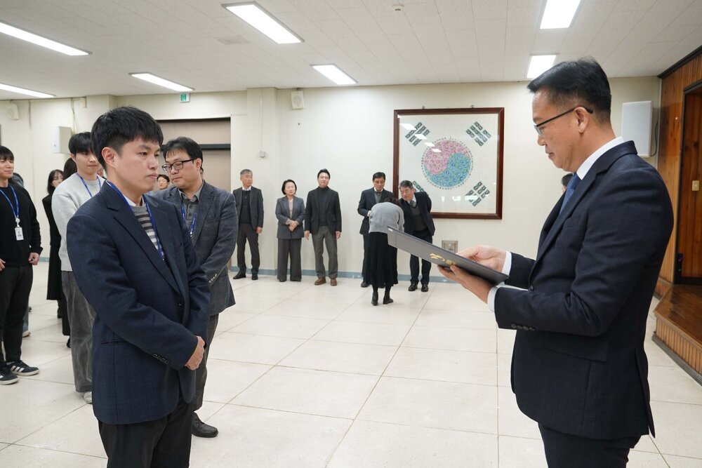 2026년 상반기 4급이하 정기인사로 정장을 입은 남성이 실내 행사장에서 한 남성에게 상장을 전달하며 내용을 읽고 있고, 남성은 남색 체크무늬 정장 자켓을 입고 두 손을 모은 상태로 서 있으며 뒤쪽으로 참석자들이 줄지어 서서 행사를 지켜보고 있다.