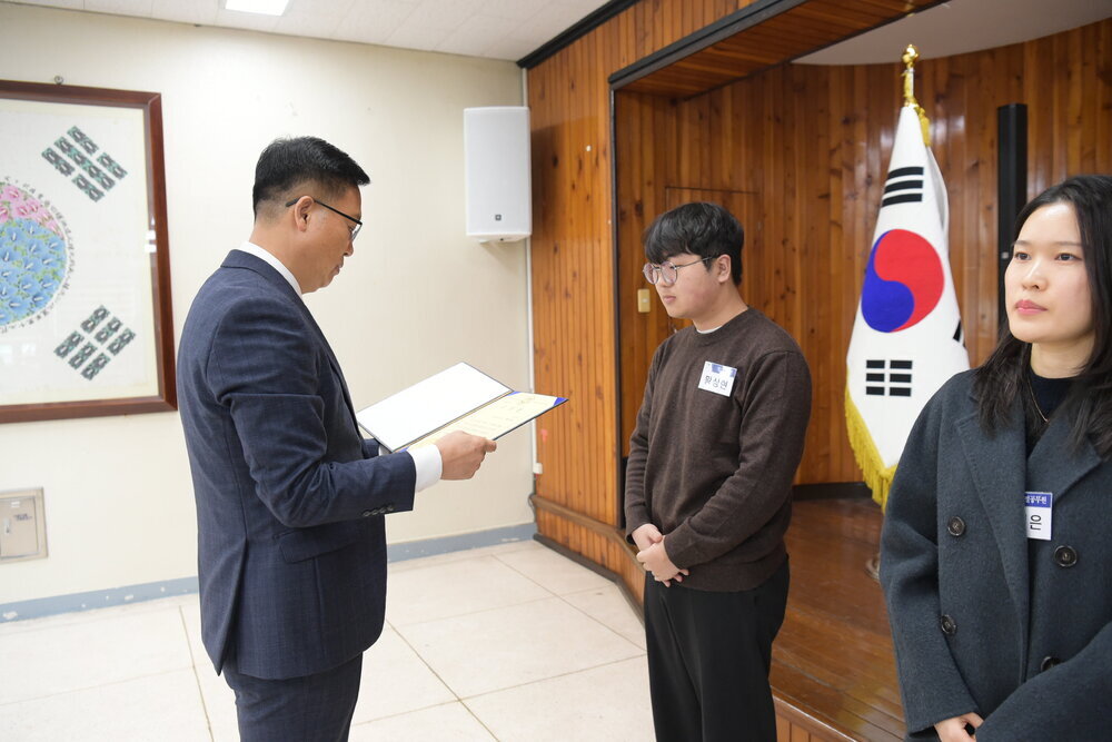 3월 정례조회에 정장을 입고 안경을 낀 남성이 검은바지에 짙은갈색 스웨터를 입은 황상현에게 패를 수여하고 있다