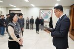2026년 상반기 4급이하 정기인사로 정장을 입은 남성이 실내 행사장에서 한 여성에게 상장을 전달하며 내용을 읽고 있고, 여성은 베이지색 목폴라 위에 기하학 무늬 베스트를 입고 사원증을 착용한 채 두 손을 모은 상태로 서 있으며 뒤쪽으로 참석자들이 줄지어 서서 행사를 지켜보고 있다.