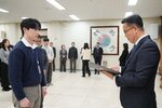 2026년 상반기 4급이하 정기인사로 정장을 입은 남성이 실내 행사장에서 한 남성에게 상장을 전달하며 내용을 읽고 있고, 남성은 흰 셔츠에 남색 니트를 겹쳐 입고 사원증을 착용한 채 두 손을 내린 상태로 서 있으며 뒤쪽으로 참석자들이 줄지어 서서 행사를 지켜보고 있다.