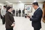 2026년 상반기 4급이하 정기인사로 정장을 입고 안경을 쓴 남성이 실내 행사장에서 한 여성에게 상장을 전달하며 내용을 읽고 있고, 여성은 갈색 무스탕 코트를 입고 두 손을 모은 채 앞에 서 있으며 뒤쪽에는 여러 사람들이 줄지어 서서 행사를 지켜보고 있다.