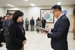2026년 상반기 4급이하 정기인사로 정장을 입고 안경을 쓴 남성이 실내 행사장에서 한 여성에게 상장을 전달하며 내용을 읽고 있고, 여성은 검은 자켓과 안경을 착용한 채 두 손을 모아 서 있으며 뒤쪽에는 여러 사람들이 줄지어 서서 행사를 지켜보고 있다.